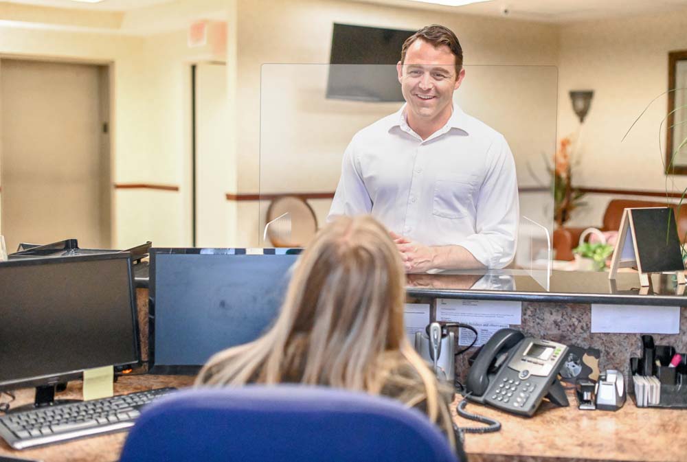 Handsome man in his 30’s stands at a office reception desk and speaks to a female receptionist. She is sitting at her desk and the man may be a client, customer, or a socializing co-worker. Office setting