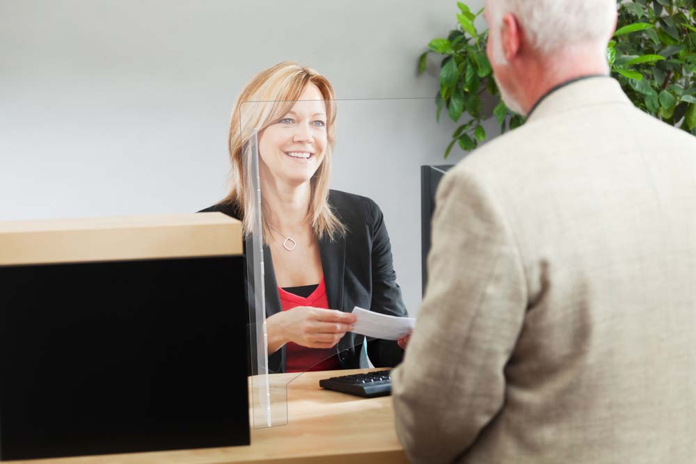 An adult man bank customer making a financial transaction with a bank teller over the counter in a retail bank. The woman caucasian bank teller is smiling and cheerfully providing the customer service. Photographed behind the shoulder of the customer in a horizontal format.